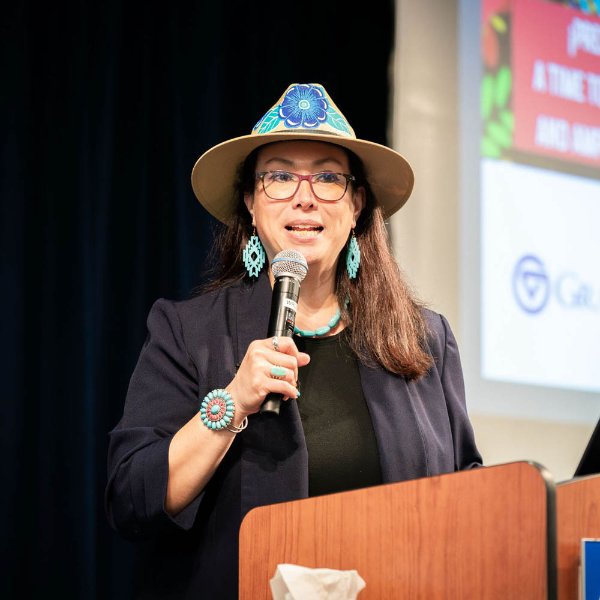Martha Villegas Miranda at a podium with microphone, wearing a hat