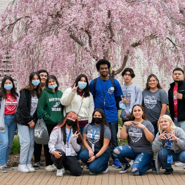 group of high school students, standing with four seated in front, standing outside on sidewalk in front of blossoming tree with pink flowers