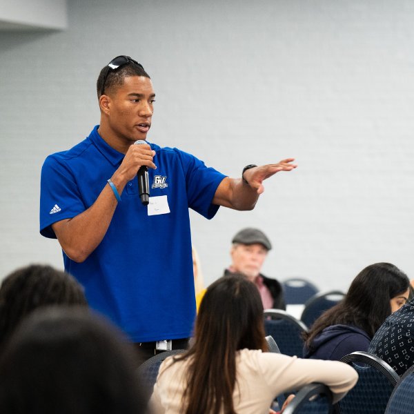 A student stands at a table and speaks into a microphone.