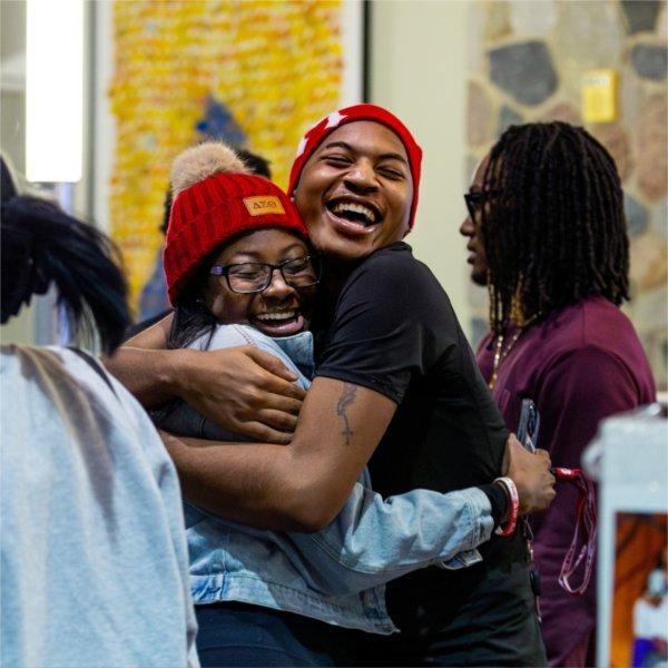 Two people wearing red winter hats embrace while having big smiles on their faces.