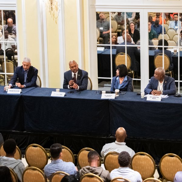 four people seated at table in front of audience; mirrored doors behind them