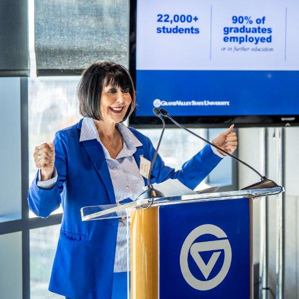 A person standing a podium smiles while gesturing with their hands. The screen behind them contains statistics saying 22,000+ students and 90% of graduates employed or in further education.
