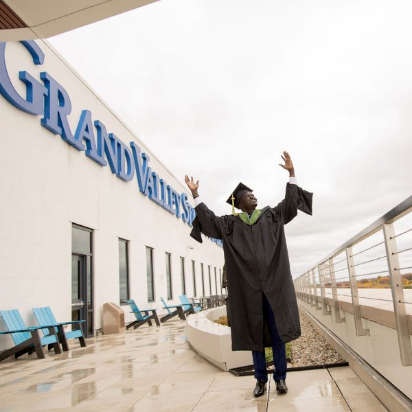 A person wearing a cap and gown has their arms raised. The words Grand Valley State are on a building in the background.
