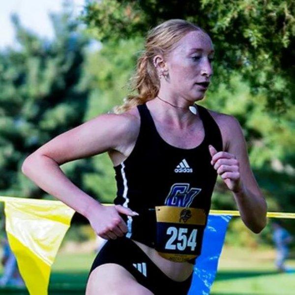 A female cross country runner in a black GVSU uniform races along a grassy course, passing colorful blue and yellow flags.