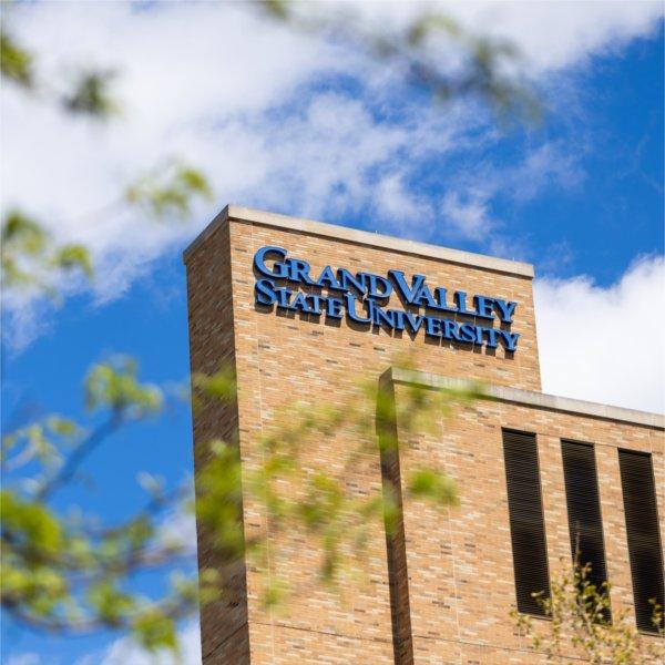 Grand Valley State University sign mounted on a brick campus building beneath a bright blue sky with scattered clouds, framed by blurred tree branches in the foreground.