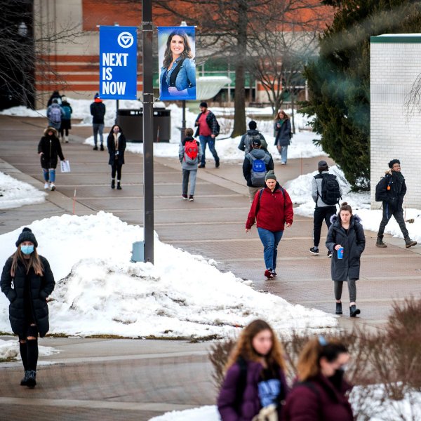 students walk on sidewalk, snow on grass areas; banner on lightpost reads Next is Now
