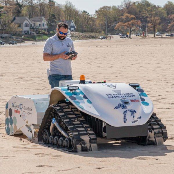 A man wearing sunglasses and a gray polo shirt stands on a sandy beach while operating a remote control for a small, tracked beach-cleaning robot.
