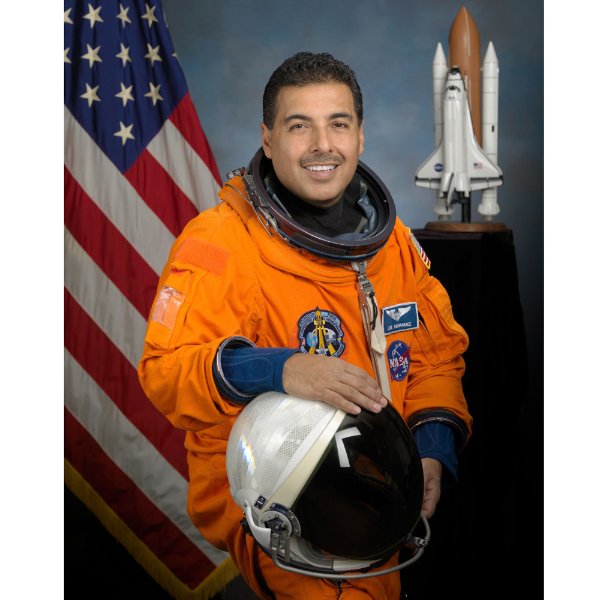 A person wearing an orange jumpsuit and holding an astronaut helmet smiles while sitting. An American flag and a rocket are in the background.