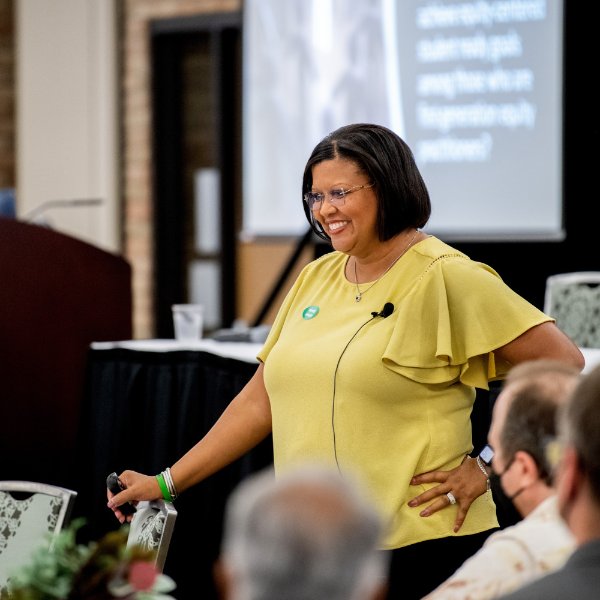Tia&#x20;Brown&#x20;McNair&#x20;stands&#x20;in&#x20;front&#x20;of&#x20;a&#x20;table&#x20;of&#x20;people,&#x20;screen&#x20;in&#x20;background&#x20;projects&#x20;her&#x20;presentation