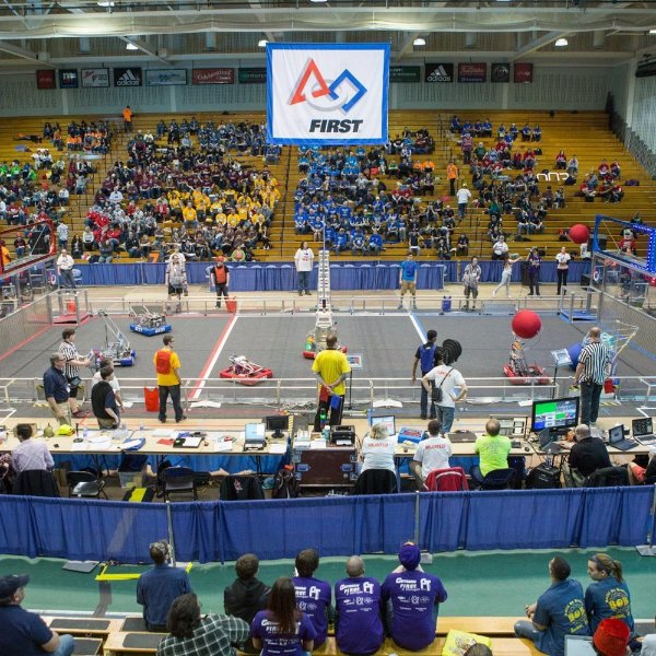 photo in the Fieldhouse of spectators in stands watching a robotics competition on the floor, banner overhead reads FIRST