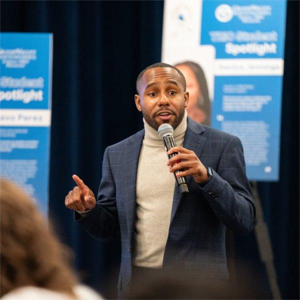 Anthony Webster with microphone, in front of posters on easels.