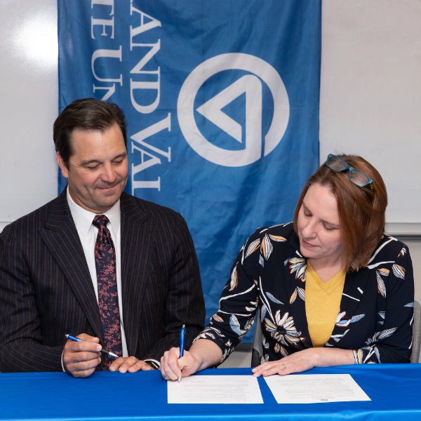 Two people are seated at a table with a blue table cloth. One person signs paper while another looks on. A Grand Valley State University flag hangs behind them.