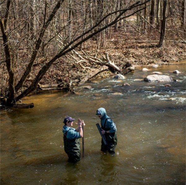 One person gestures to another while both are standing in a stream.