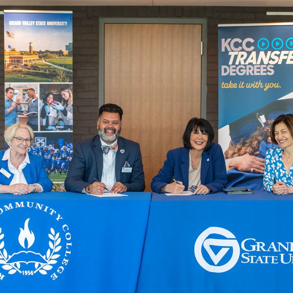 four people seated at two tables, at left is Kellogg Community College leaders with banner in back, at right are President Mantella and Provost Mili with banners in background