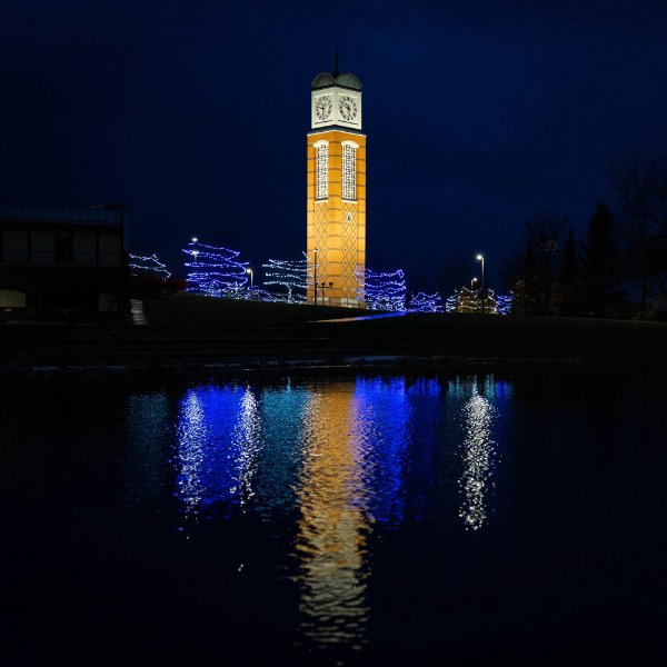 Cook Carillon Tower lit up with trees lit with blue lights surrounding it; tower is reflected in Zumberge pond.