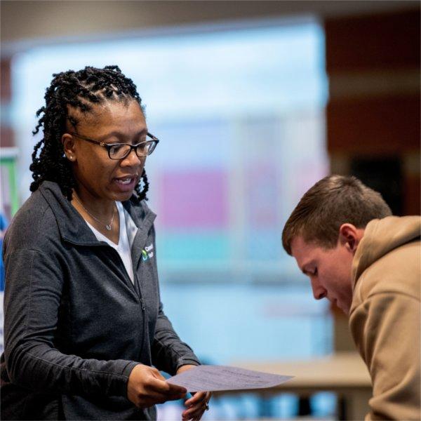 A person holding a paper speaks to a person who is bending down toward the table.