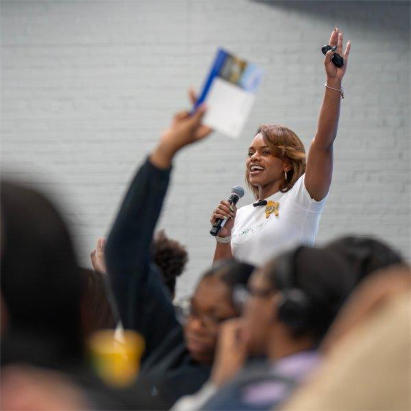speaker in white polo shirt with left arm raised before a group of people seated at tables