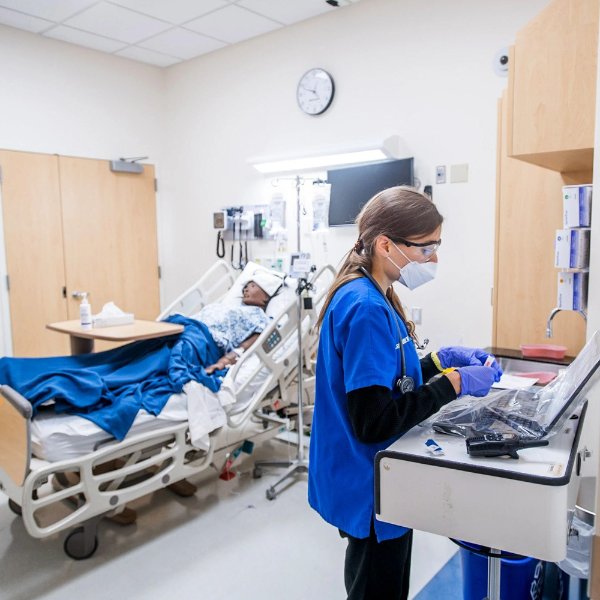 a student in blue scrub shirt looks at a computer monitor in a simulated hospital room, with a mannequin on the bed, in the Sim Center in the DeVos Center for Interprofessional Health