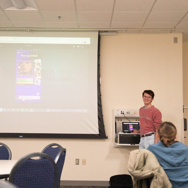 Caleb White stands to the left of a projection screen that shows a Spotify screenshot of Taylor Swift.