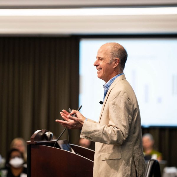 James Lang holds out hands while speaking from the podium, he is dressed in a tan suit