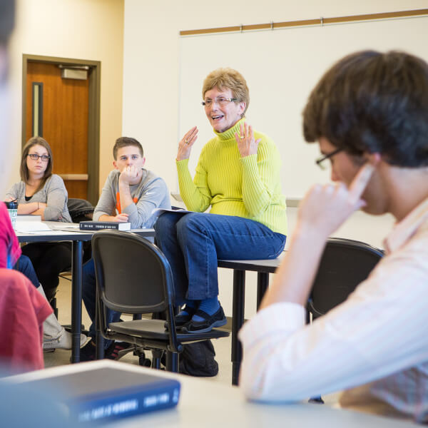 woman sitting on desk, talking with students in class