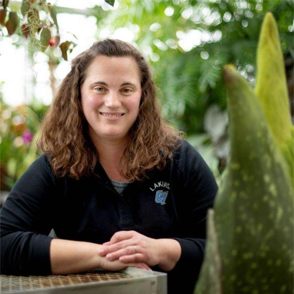 A person with arms folded on table smiles while standing next to a plant.