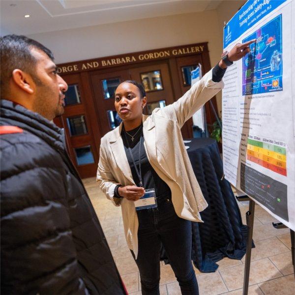 A person stands beside a large research poster, pointing to a section of it while speaking to another attendee.