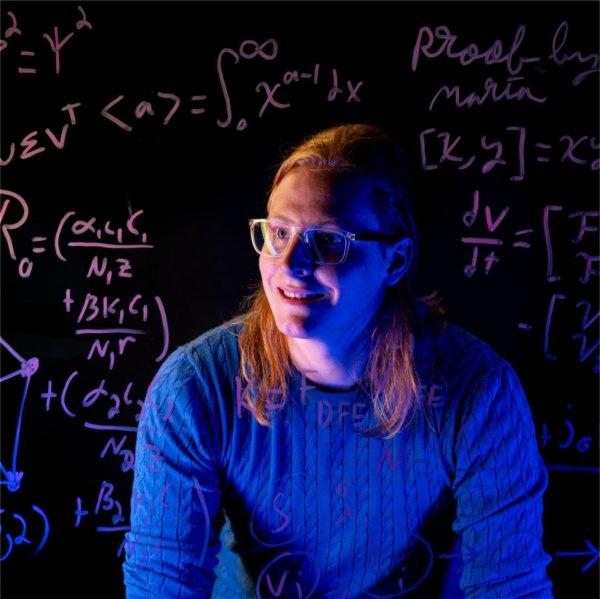 A student wearing glasses and a blue sweater sits behind a transparent board covered with handwritten mathematical equations, formulas and diagrams.