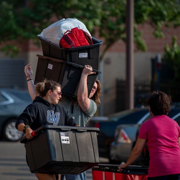 A person holds a plastic bin while another standing behind the person hoists two plastic bins of belongings.