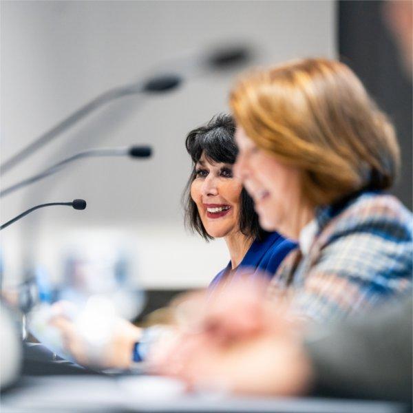 One person smiles as another speaks. Microphones are before them.