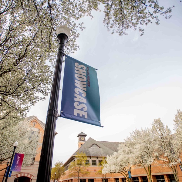 A Reach Higher Showcase banner hangs from a post outside the DeVos Center, surrounded by blossoming trees.
