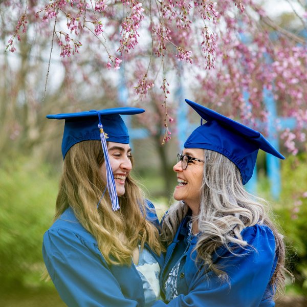 Two people wearing caps and gowns look at each other and are smiling. The blossoms from a tree hang overhead.