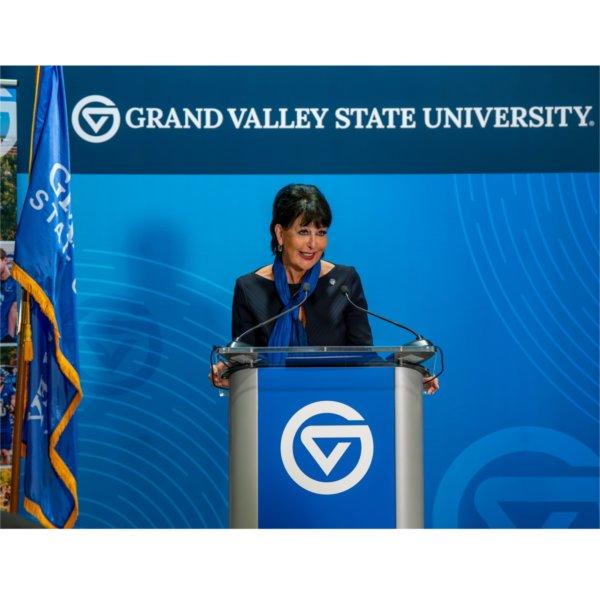 A person smiles while standing at a podium. The words Grand Valley State University are on a banner in the background. A flag is also in the background.