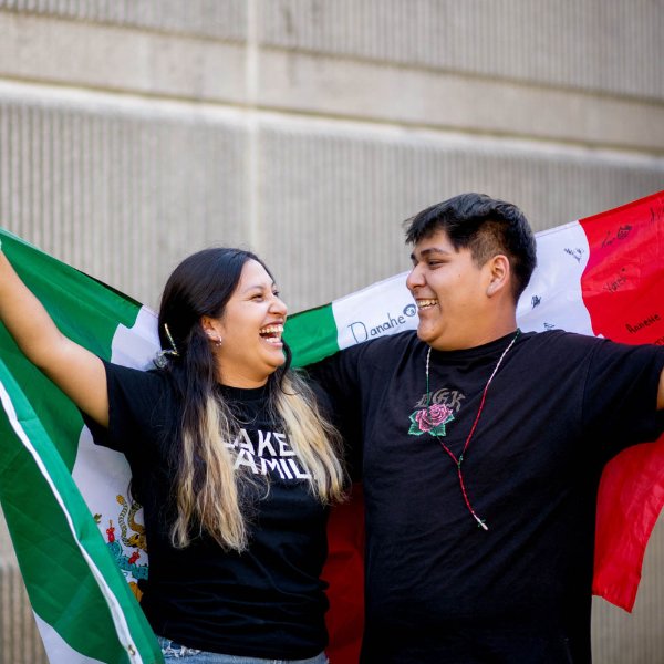 Two students, Jackelyn Palmas and Jose Medina, stand together laughing and holding Mexican flags.