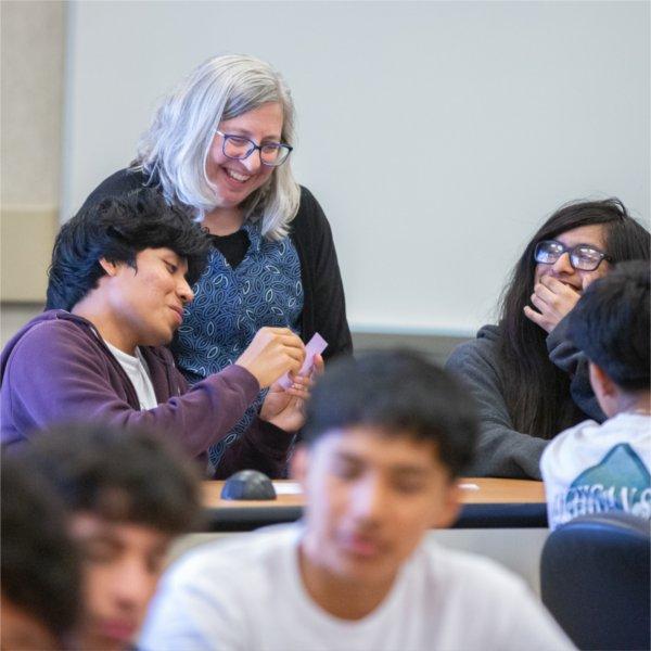 a young student shows a card to a woman leaning over his shoulder, while a classmate laughs and looks on