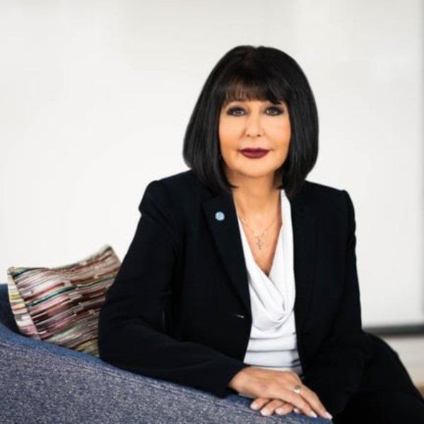 A person poses for a photo with hands folded on the arm of a chair.
