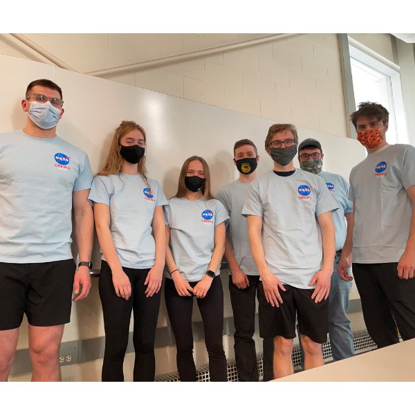 Seven students in NASA crew t-shirts standing against a wall and wearing face masks.