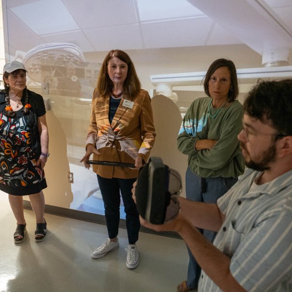 four people in immersive room with screens showing operating room on walls, man in front holds a VR headset