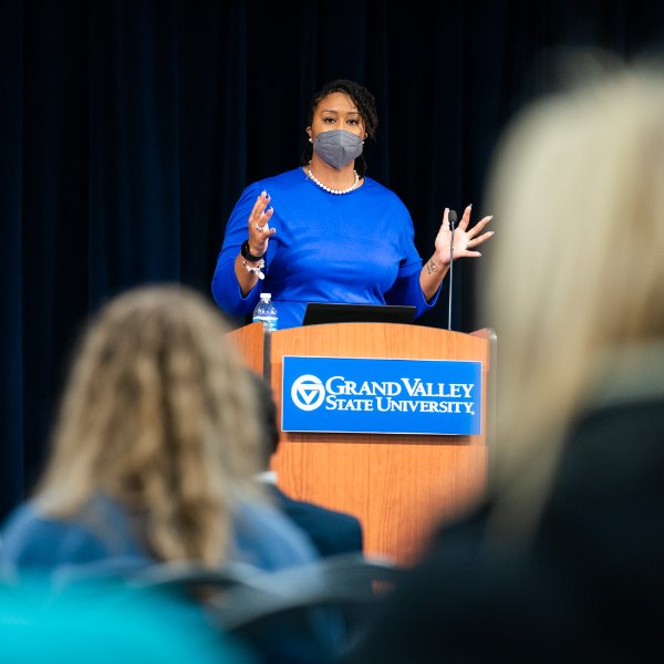 Littisha Bates speaks from behind a podium, using her hands, several people in audience seated in front.
