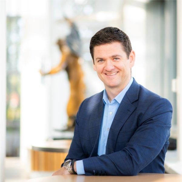 Milo&#353; Topi&#263; with arm resting on table, sculpture behind him, he is dressed in blue suit jacket and light blue shirt