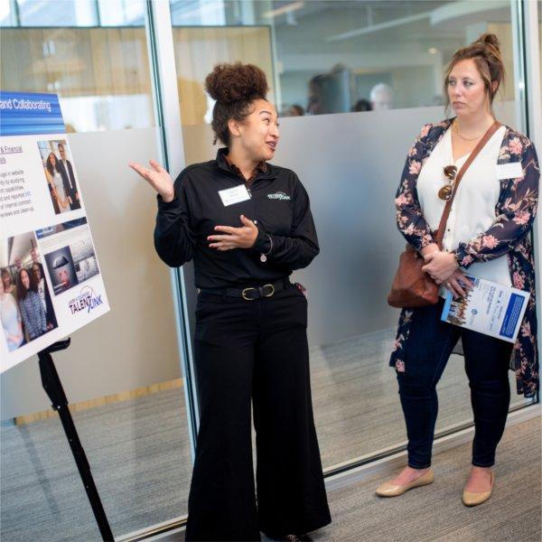 A person gestures toward a poster board while talking to others.