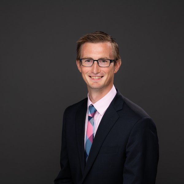 Zachary DeBruine seated for a headshot in a dark suit, colorful tie