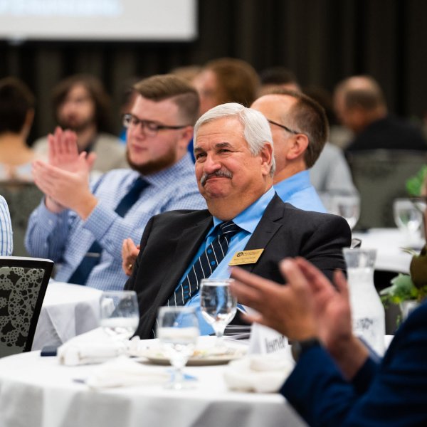 Paul Plotkowski is seated at a table smiling as others clap for his accomplishments