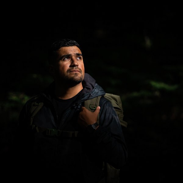 A person stands in the woods holding a backpack as the light hits their face.