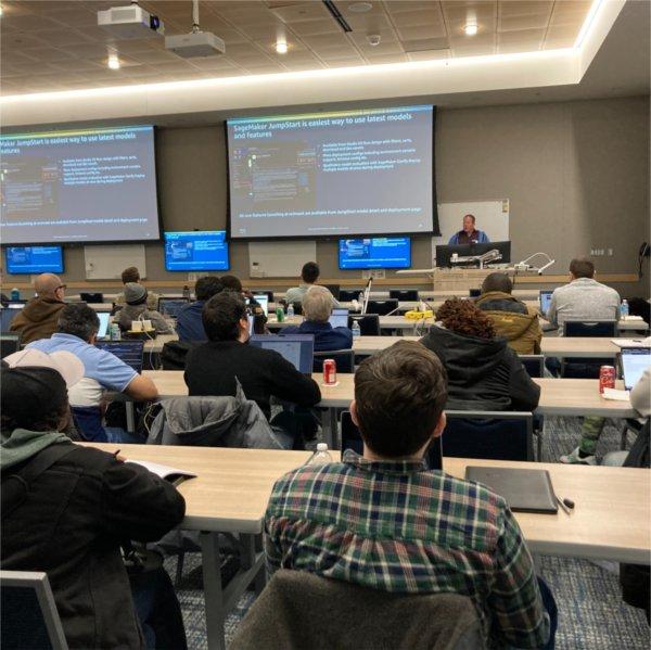 rows of people seated at long tables in classroom, person at front with projected screens behind, about coding and web development