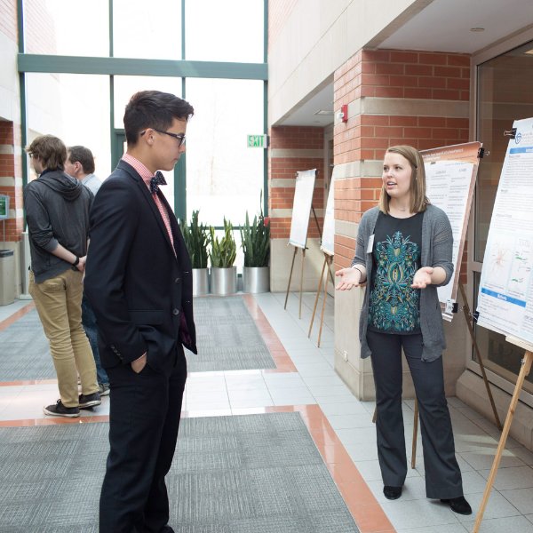 A Student Scholars Day participant stands next to a poster explaining it to another person.