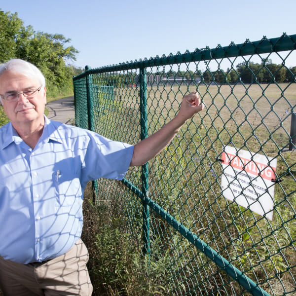 Rick Rediske stand next to the site of the former Wolverine World Wide tannery in Rockford, Michigan. He holds a chain link fence with a sign warning of environmental contamination.