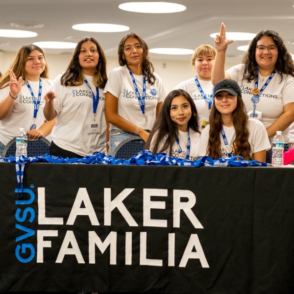 students seated and standing by a table with a drape that says GVSU Laker Familia, they are all wearing white t-shirts