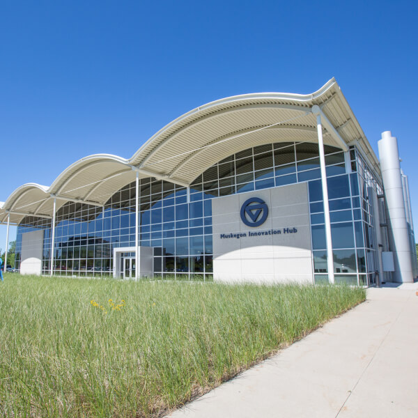 A photo shows the Muskegon Innovation Hub from the front, with large arching glass windows and a front landscape of dune grass.