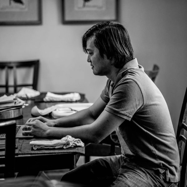 a black and white photo of John Wen typing on a laptop at his dining room table; he is wearing a golf shirt